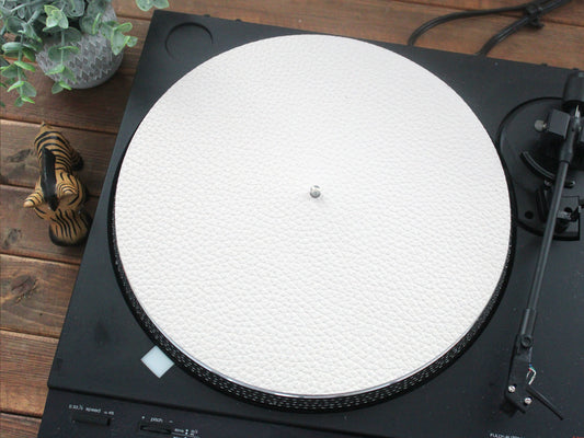 A white leather turntable mat placed on a turntable, with a record player arm and a plant in the background.