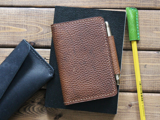 A brown full-grain leather folio pocket journal wallet is displayed on top of a black notebook, with a green and a yellow pen beside it.