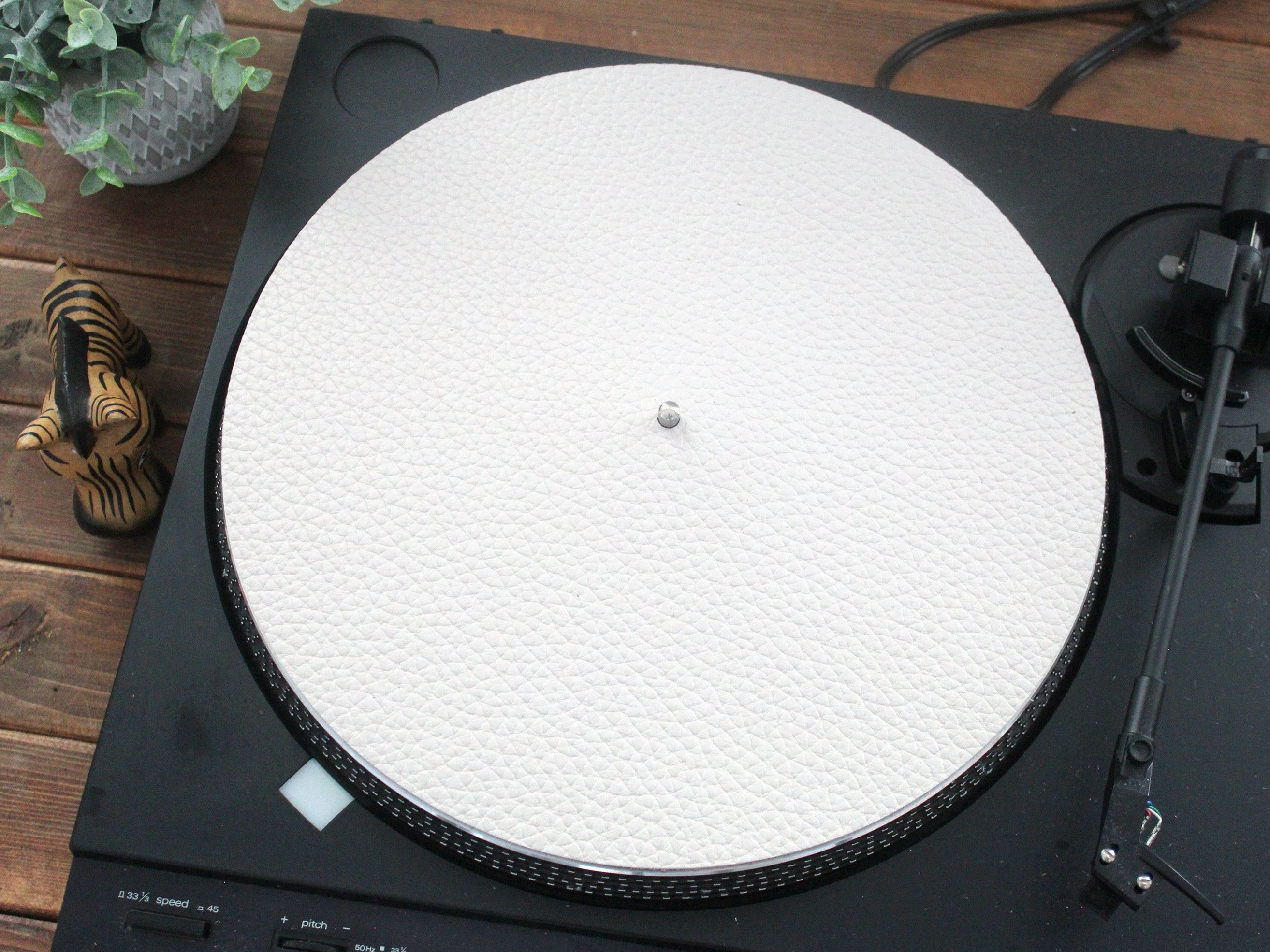 A white leather turntable mat placed on a turntable, with a record player arm and a plant in the background.
